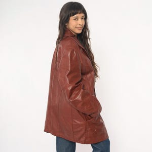May include: A vintage, oversized, reddish-brown leather jacket with a classic collar and two front pockets. The jacket is worn over blue jeans. The leather has a slightly glossy finish. The jacket appears to be in good condition.