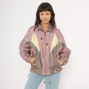 May include: A vintage mauve windbreaker jacket with a zip-up front, featuring a colour-block design with cream and grey panels. The jacket has a drawstring waist, pockets, and floral patterned accents. The model is wearing light blue jeans.