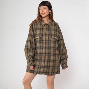 May include: A long-sleeved, oversized plaid shirt in shades of brown and green. The shirt features a button-down front, two chest pockets, and buttoned cuffs. The fabric has a soft, slightly worn appearance, suggesting a vintage or relaxed style.