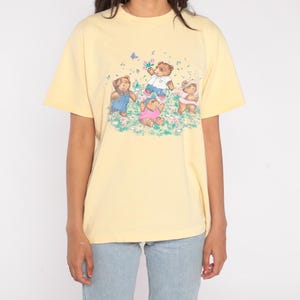 May include: A light yellow t-shirt featuring a graphic of four teddy bears in a field of flowers. The bears are wearing different outfits, including overalls and a pink shirt. A butterfly is also present in the design.