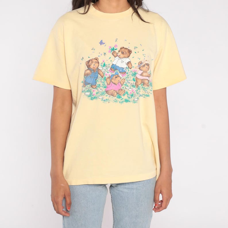 May include: A light yellow t-shirt featuring a graphic of four teddy bears in a field of flowers. The bears are wearing different outfits, including overalls and a pink shirt. A butterfly is also present in the design.