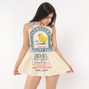 May include: A sleeveless cream-colored mini dress featuring a graphic print resembling a vintage feed sack. The design includes a yellow chick, blue text reading FARM GROWN QUALITY FEED, and red text stating MANUFACTURED BY BAUER FEED & HATCHERY.