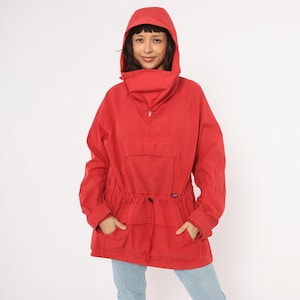May include: A red hooded anorak jacket with a drawstring waist and a front pocket. The jacket has a half-zip closure and a high collar. The model is wearing blue jeans.