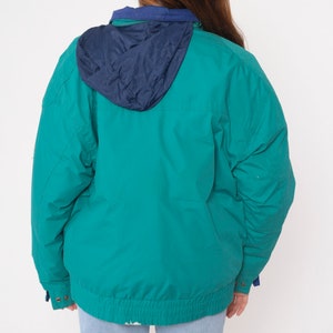 K&ouml;nnte beinhalten: Eine tealblaue Kapuzenjacke mit einem marineblauen Futter. Die Jacke hat einen Rei&szlig;verschluss und eine Kapuze mit Kordelzug.