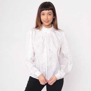 May include: White long-sleeve blouse with a high neckline and a subtle floral pattern. The blouse has a gathered front and buttoned cuffs. The fabric appears to be a lightweight material.