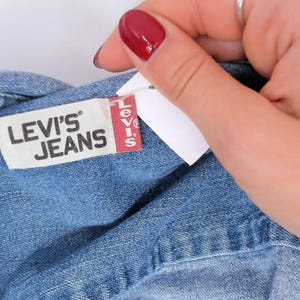 K&ouml;nnte beinhalten: Nahaufnahme eines blauen Jeanskleidungsst&uuml;cks mit einem Etikett mit der Aufschrift "LEVI'S JEANS" in wei&szlig;er Schrift auf wei&szlig;em Hintergrund. Das Levi's-Etikett hat einen roten vertikalen Streifen mit dem Levi's-Logo. Eine Hand mit rotem Nagellack h&auml;lt das Kleidungsst&uuml;ck.