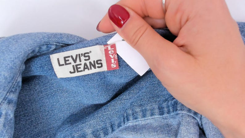 K&ouml;nnte beinhalten: Nahaufnahme eines blauen Jeanskleidungsst&uuml;cks mit einem Etikett mit der Aufschrift "LEVI'S JEANS" in wei&szlig;er Schrift auf wei&szlig;em Hintergrund. Das Levi's-Etikett hat einen roten vertikalen Streifen mit dem Levi's-Logo. Eine Hand mit rotem Nagellack h&auml;lt das Kleidungsst&uuml;ck.