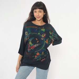 May include: Dark navy blue long-sleeve shirt with a colorful abstract graphic design. The design features a scooter, musical instruments, and various shapes in red, green, and yellow. The shirt is a loose fit.