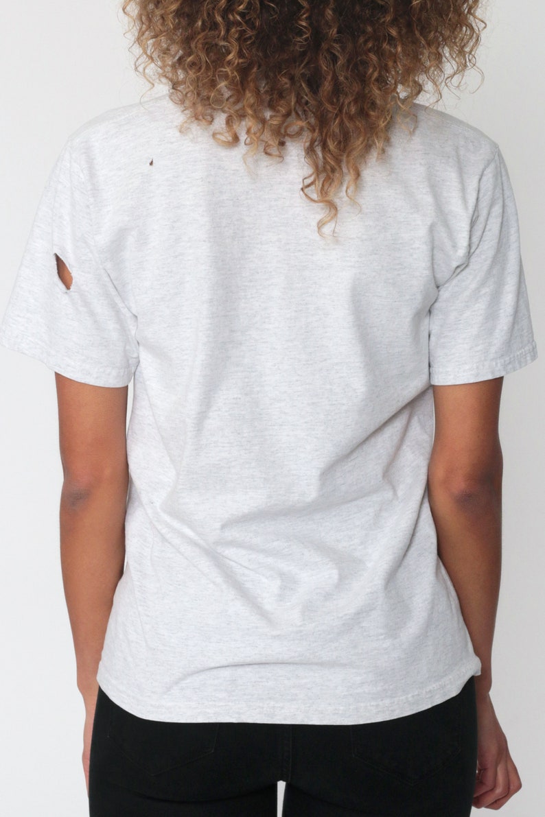 May include: A gray short-sleeved t-shirt with a distressed look. The shirt has a small hole near the shoulder seam.