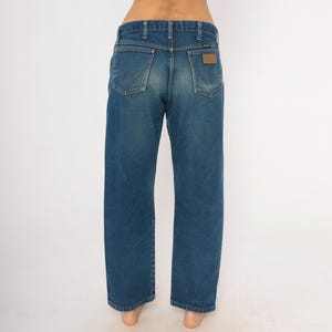 May include: Blue denim jeans with a classic straight leg fit. The jeans feature the Wrangler logo on the back pockets and a brown leather patch. The jeans are a medium wash and have a traditional five-pocket design.
