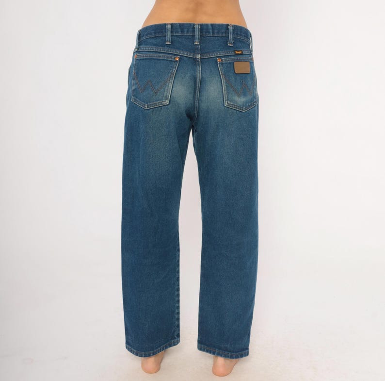 May include: Blue denim jeans with a classic straight leg fit. The jeans feature the Wrangler logo on the back pockets and a brown leather patch. The jeans are a medium wash and have a traditional five-pocket design.