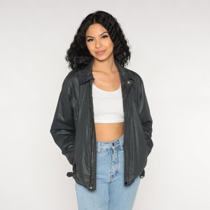 May include: Dark gray jacket with a collared neckline and a zipper closure. The jacket has two front pockets and a belt detail on the sides. The model is wearing a white crop top and light blue jeans.