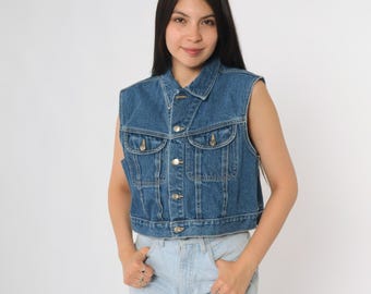 May include: A sleeveless denim vest in a medium blue wash. The vest features a button-up front, two chest pockets, and a classic collar. The vest is a cropped style.