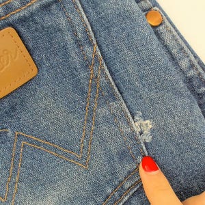 May include: Close-up of blue denim jeans with a brown leather "Wrangler" patch. The jeans have a distressed area near the pocket and a copper-colored button. The stitching is a contrasting tan color.