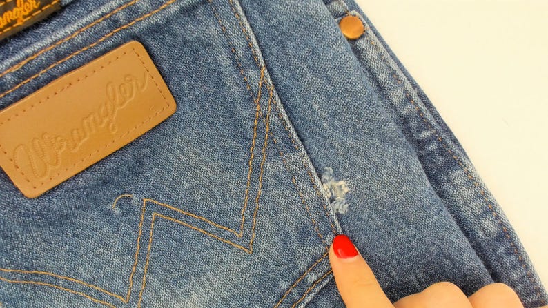 May include: Close-up of blue denim jeans with a brown leather "Wrangler" patch. The jeans have a distressed area near the pocket and a copper-colored button. The stitching is a contrasting tan color.