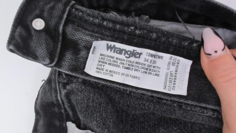 May include: Close-up of black Wrangler jeans. The label reads "Wrangler 13MWZWK 34 x 30". The jeans are made of 100% cotton and made in Mexico of US fabric. The care instructions are also visible on the label.
