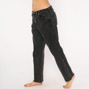 May include: Black denim jeans with a classic five-pocket design. The jeans have a straight leg cut and a faded appearance. The dark wash denim features copper-colored rivets and a button closure. The jeans are worn casually.