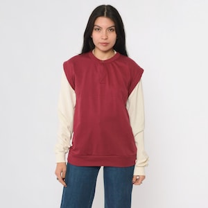 Peut inclure: Un pull-over bordeaux avec une chemise à manches longues crème en dessous. Le pull-over a une encolure boutonnée et une coupe décontractée. Le modèle porte un jean bleu. La tenue est décontractée et confortable.