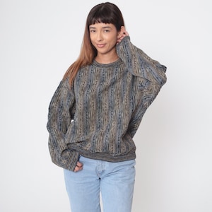 May include: A textured, oversized sweater in shades of blue and tan. The sweater has a ribbed knit pattern and a crew neck. The sleeves are long and the cuffs and hem are ribbed. The person is wearing light blue jeans.
