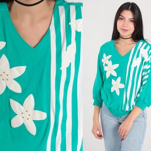 May include: A teal and white striped sweater with white flower appliques. The sweater has a v-neck and long sleeves.