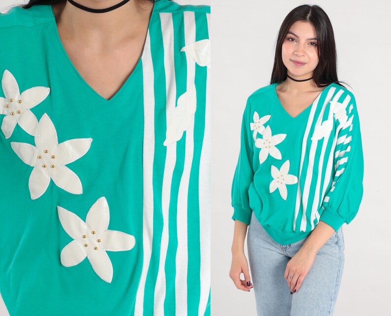 May include: A teal and white striped sweater with white flower appliques. The sweater has a v-neck and long sleeves.