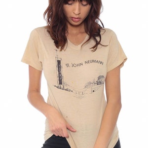 May include: Beige v-neck t-shirt featuring a black and white illustration of a tall structure with the text "ST. JOHN NEUMANN". The shirt is made of a soft, lightweight material.