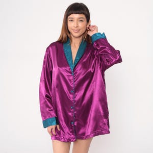 May include: A long-sleeved, button-down satin shirt in a vibrant purple hue, accented with teal trim on the collar and cuffs. The shirt has a relaxed fit and a glossy sheen, suggesting a vintage or retro style.