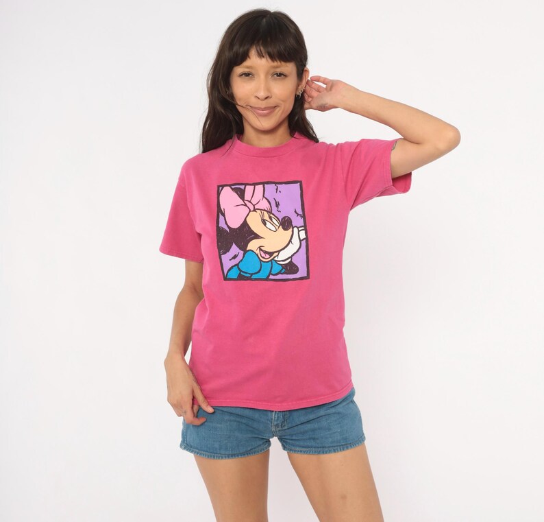 May include: A short-sleeved, crew-neck t-shirt in a vibrant magenta pink colour. The centre of the shirt features a square graphic print of a cartoon mouse character with a pink bow and blue dress, set against a light purple background.