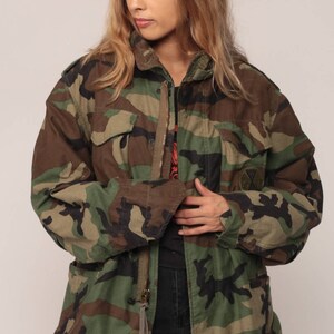 May include: A camouflage field jacket featuring a woodland pattern in shades of green, brown, and black. The jacket includes a front metal zipper closure, two chest pockets with buttoned flaps, and a circular patch on the left chest area.