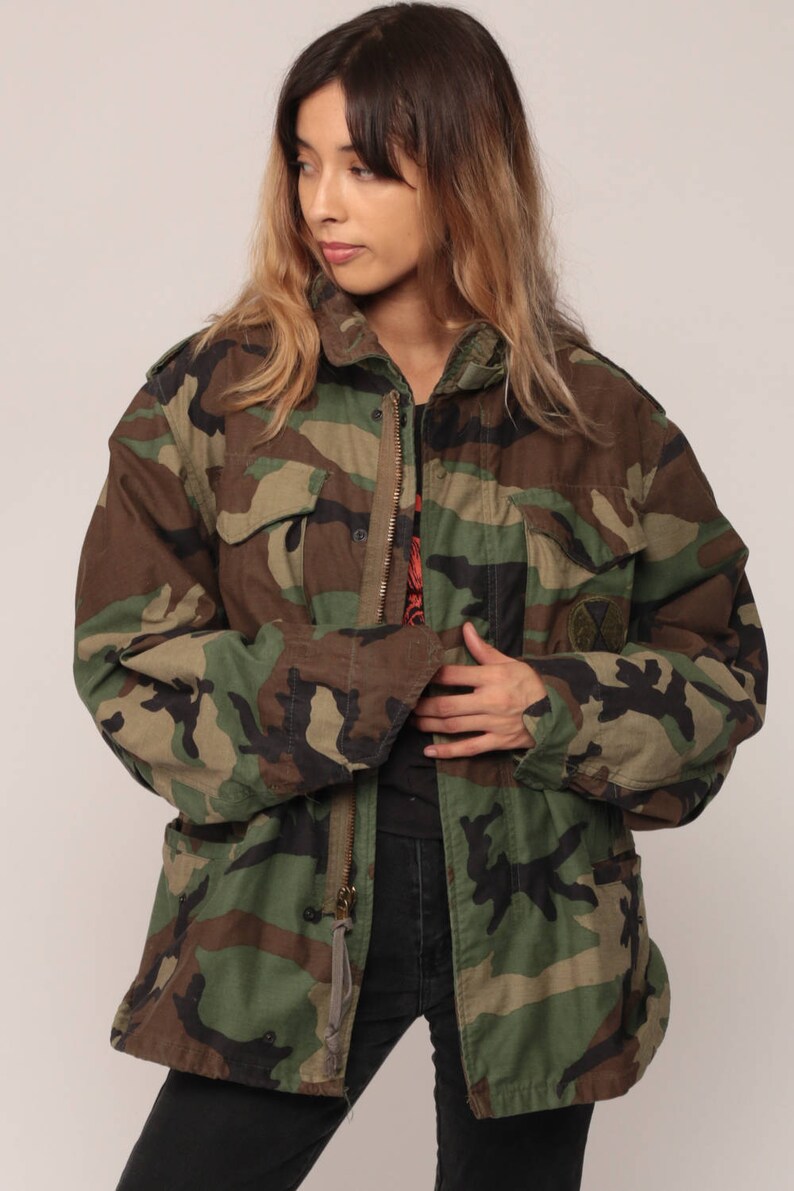 May include: A camouflage field jacket featuring a woodland pattern in shades of green, brown, and black. The jacket includes a front metal zipper closure, two chest pockets with buttoned flaps, and a circular patch on the left chest area.