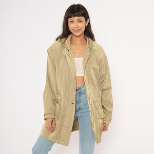May include: A long, gold-colored coat with a hood. The coat has a button closure and two front pockets. The model is wearing blue jeans and a white top. The coat appears to be made of a shiny, lightweight material.