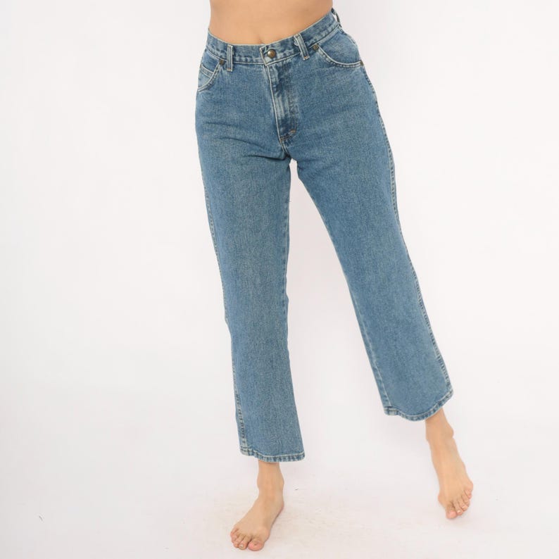 May include: Light blue denim jeans with a classic five-pocket design. The jeans have a straight leg cut and a button and zipper closure. The denim is a medium wash, and the jeans appear to be vintage.