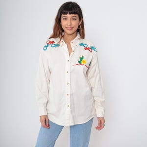 May include: Cream-colored button-down shirt with long sleeves. The shirt features embroidered designs of lizards in red and blue on the shoulders, and a cactus and sun on the front. The model is wearing light blue jeans.