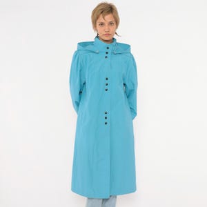 May include: A bright turquoise long-length coat featuring a hood, long sleeves, and a front placket with a series of dark circular snap buttons. The coat has a structured collar and a clean, solid-colored design with a straight hemline.