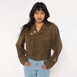 May include: An olive green military-style cropped jacket featuring a collared neckline, two front chest pockets with flaps, and long sleeves. The jacket is adorned with various small metallic pins on the chest and embroidered patches on the sleeves.