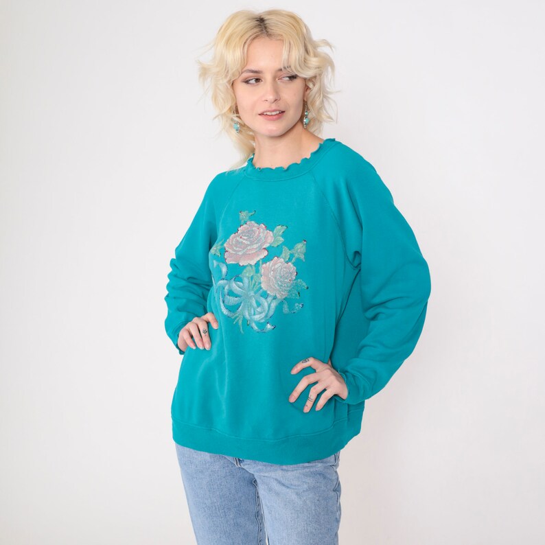 May include: A turquoise blue sweatshirt with a floral print of pink and white roses. The sweatshirt has a ruffled neckline and long sleeves.
