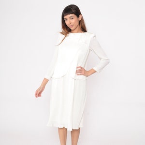 May include: A white dress with a textured fabric and a layered design. The dress has a scoop neck and long sleeves with button cuffs. The dress has a knee-length skirt with a ruffled hem.