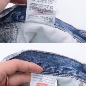 Puede incluir: Primer plano del interior de unos vaqueros azules con dos etiquetas blancas. Una etiqueta tiene instrucciones de lavado, y la otra etiqueta dice "Wrangler" e indica la talla 29 x 32 Regular Fit.