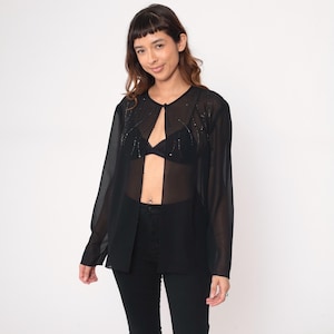 May include: A sheer black long-sleeve top with a keyhole neckline and embellished with small, sparkling beads. The top is paired with black pants. The overall style is elegant and semi-transparent.