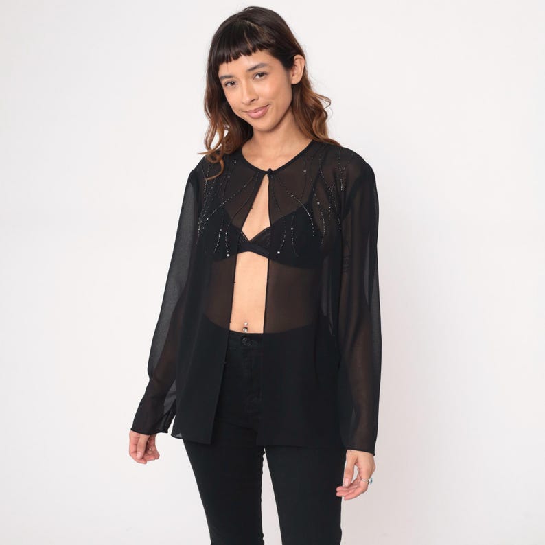 May include: A sheer black long-sleeve top with a keyhole neckline and embellished with small, sparkling beads. The top is paired with black pants. The overall style is elegant and semi-transparent.