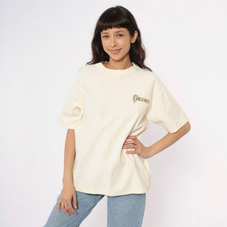 May include: Cream-colored t-shirt with short sleeves and a crew neck. The shirt features the text "GIRL SCOUT" in a decorative font on the upper chest. The model is wearing light blue jeans.