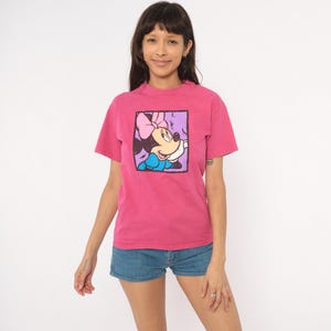 May include: A bright pink short-sleeved t-shirt featuring a square graphic print on the chest. The graphic depicts a cartoon mouse character with a pink bow, blue dress, and white gloves, framed by a thick black border against a light purple background.