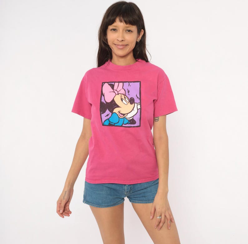 May include: A bright pink short-sleeved t-shirt featuring a square graphic print on the chest. The graphic depicts a cartoon mouse character with a pink bow, blue dress, and white gloves, framed by a thick black border against a light purple background.