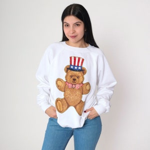 Könnte beinhalten: Weißer Pullover mit einem braunen Teddybär, der einen rot-weiß-blauen Onkel-Sam-Hut trägt. Der Bär trägt eine Fliege.