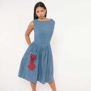 May include: A sleeveless blue midi dress featuring a round neckline and a fitted bodice. The skirt is pleated and includes a large patch pocket on the front, which is decorated with a red cat appliqué featuring white polka dots and black whiskers.