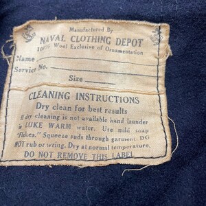 May include: A tan label with black text that reads "Manufactured By NAVAL CLOTHING DEPOT 100% Wool Exclusive of Ornamentation" and "CLEANING INSTRUCTIONS Dry clean for best results If dry cleaning is not available hand launder in LUKE WARM water. Use mild soap "Flakes." Squeeze suds through garment. DO NOT rub or wring. Dry at normal temperature, DO NOT REMOVE THIS LABEL". The label has a small anchor symbol in the top left corner and a larger anchor symbol in the top right corner.