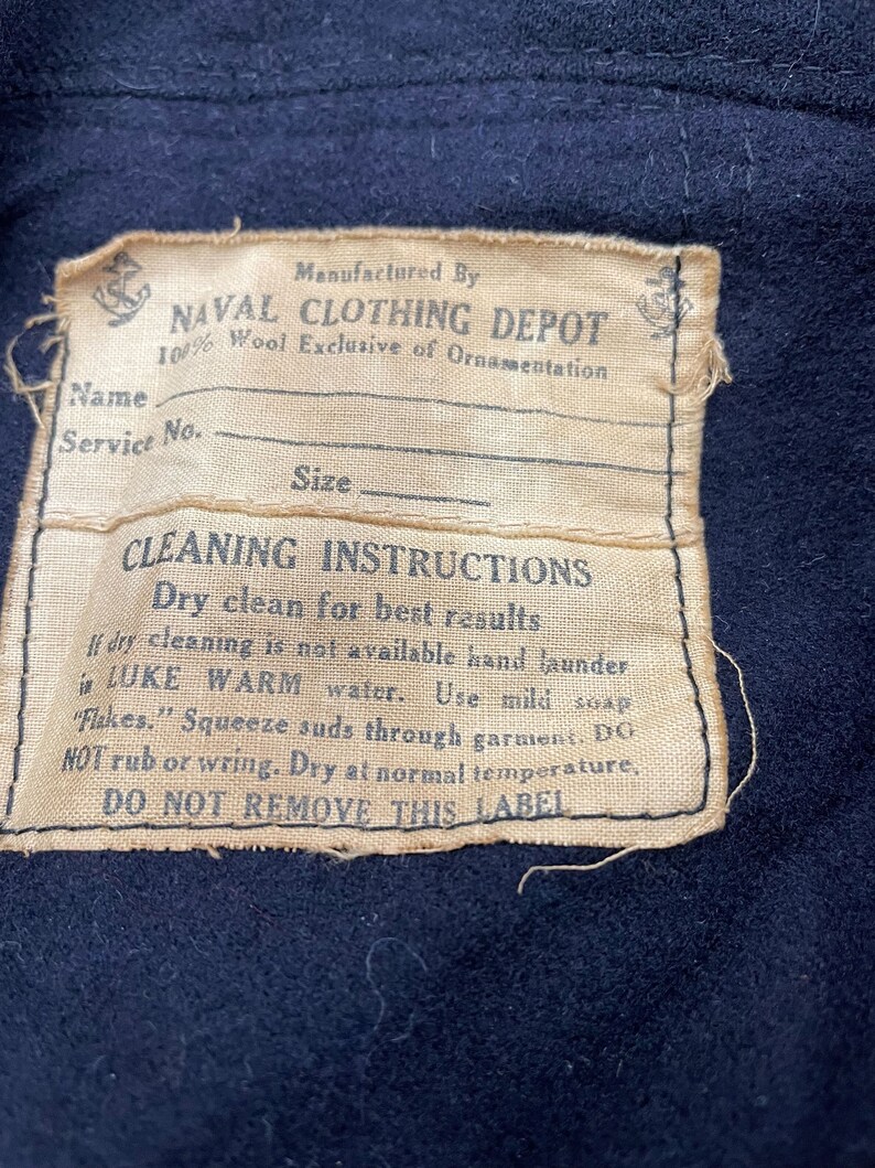 May include: A tan label with black text that reads "Manufactured By NAVAL CLOTHING DEPOT 100% Wool Exclusive of Ornamentation" and "CLEANING INSTRUCTIONS Dry clean for best results If dry cleaning is not available hand launder in LUKE WARM water. Use mild soap "Flakes." Squeeze suds through garment. DO NOT rub or wring. Dry at normal temperature, DO NOT REMOVE THIS LABEL". The label has a small anchor symbol in the top left corner and a larger anchor symbol in the top right corner.