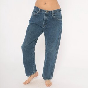 May include: Blue denim jeans with a classic five-pocket design. The jeans have a straight leg cut and a button and zipper closure. The denim is a medium wash, and the jeans appear to be vintage.