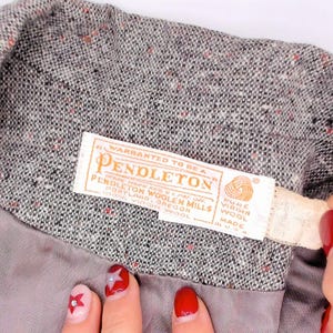 May include: Close-up of a gray tweed jacket with a Pendleton label. The label is cream-colored with the Pendleton logo and text. The jacket's fabric has flecks of red and black.