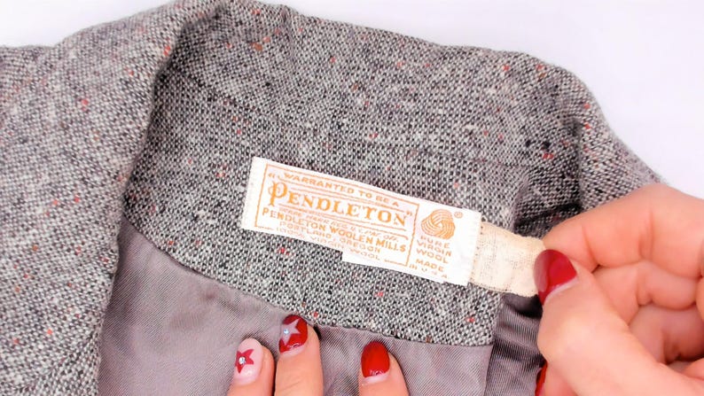 May include: Close-up of a gray tweed jacket with a Pendleton label. The label is cream-colored with the Pendleton logo and text. The jacket's fabric has flecks of red and black.